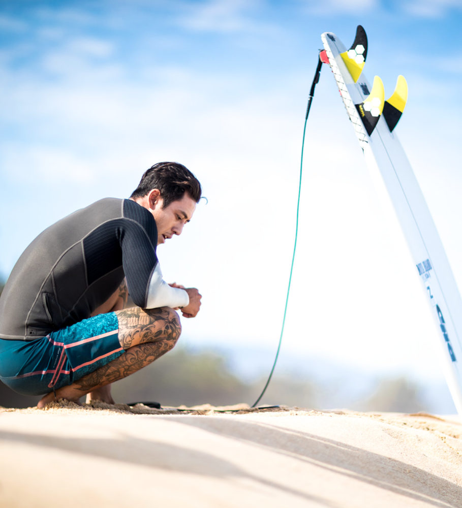 O'NEILL（オニール）チームメンバー ELI OLSON（エリ・オルソン） 砂浜にサーフボードを立てて座っている